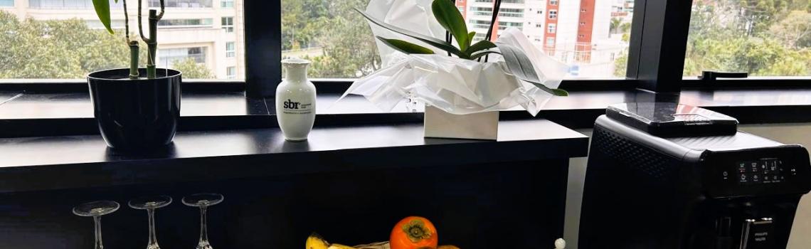 Imagem da mesa do café, na mesa tem uma cesta com frutas ao centro, canecas e taças no lado esquerdo da foto, cafeteira com um açucareiro do lado direito da foto. E vasos ao fundo na janela.  